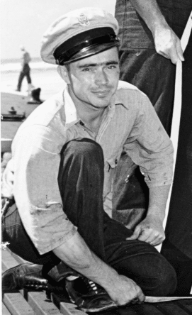 Black and white image of a young Gunner's Mate, Paul G. Saunders, kneeling in uniform on a dock.